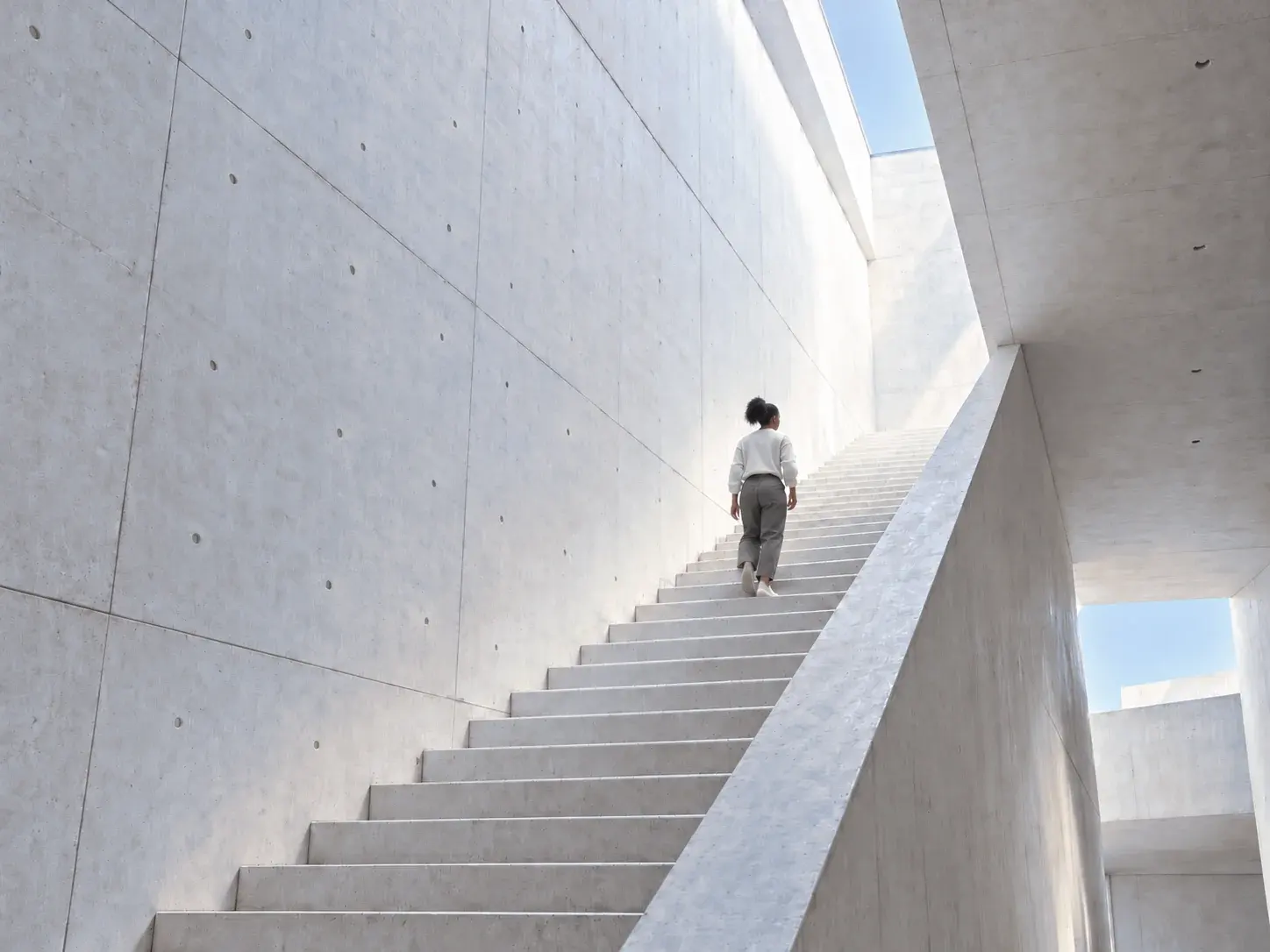 Une jeune femme monte un grand escalier de béton baigné de lumière.